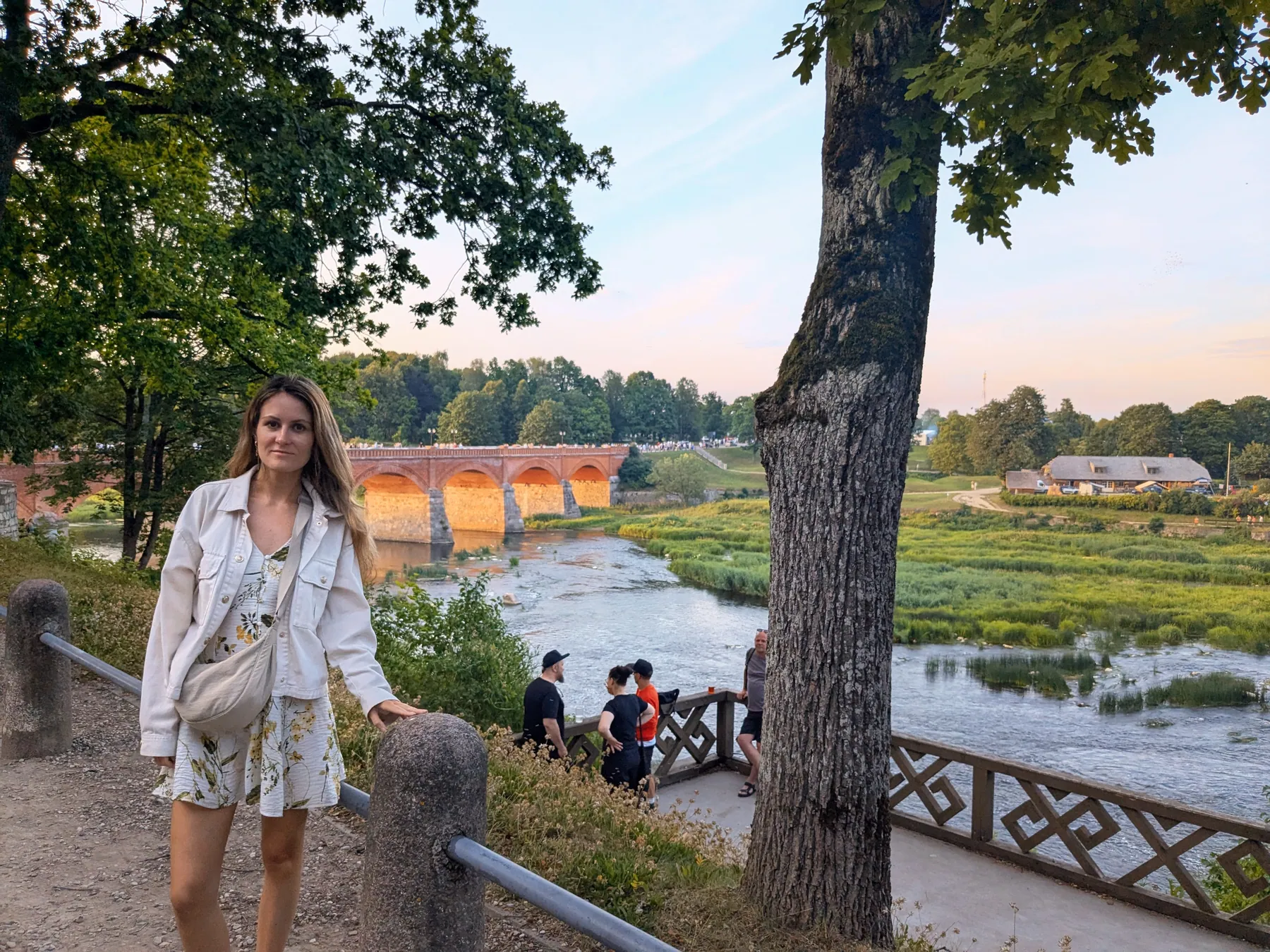 A traveler beside the Venta River and Kuldīga brick bridge at golden hour