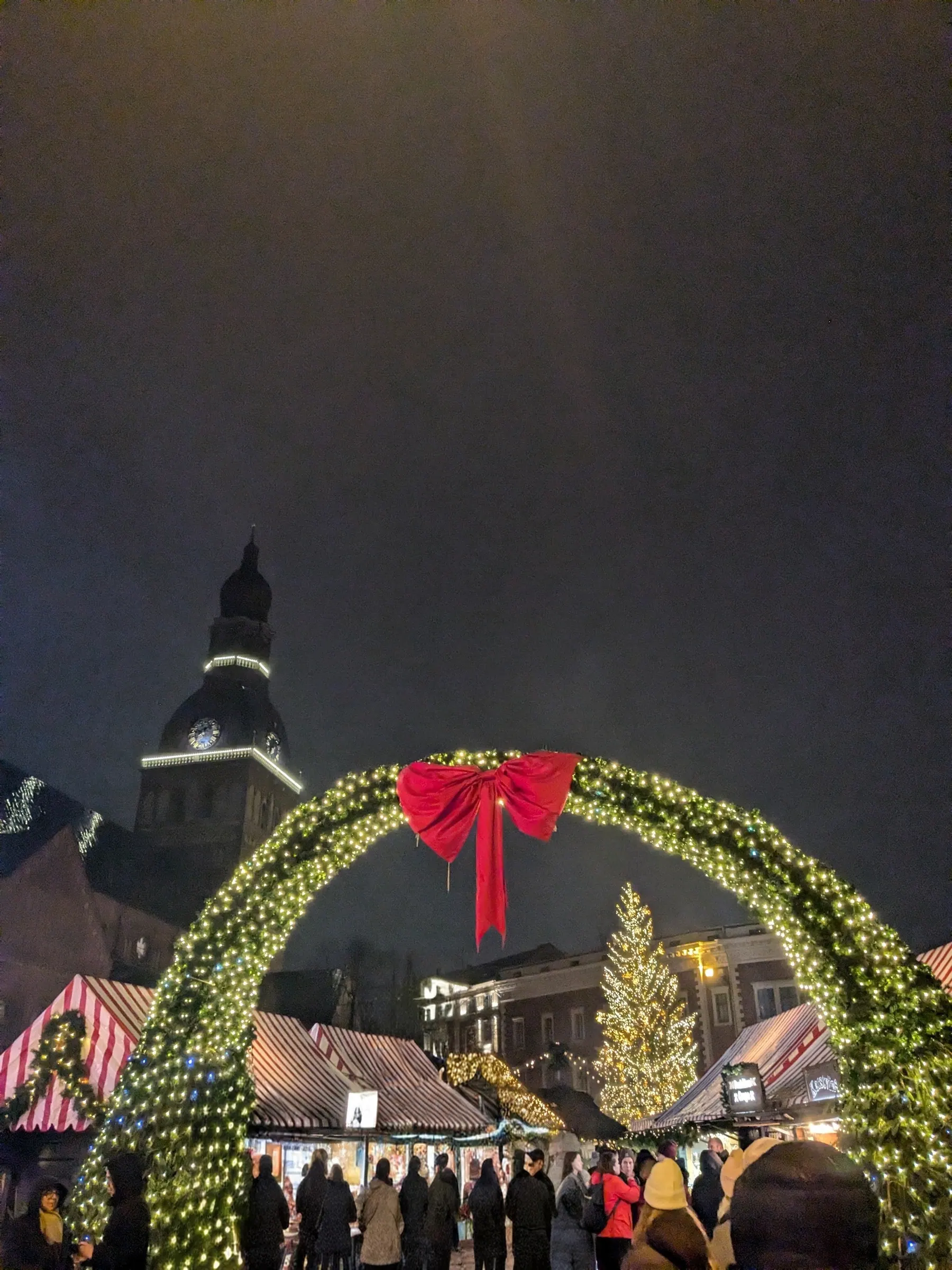 Lighted pine archway with a large red ribbon bow framing Dome Square Christmas Market and Riga Cathedral spire at night