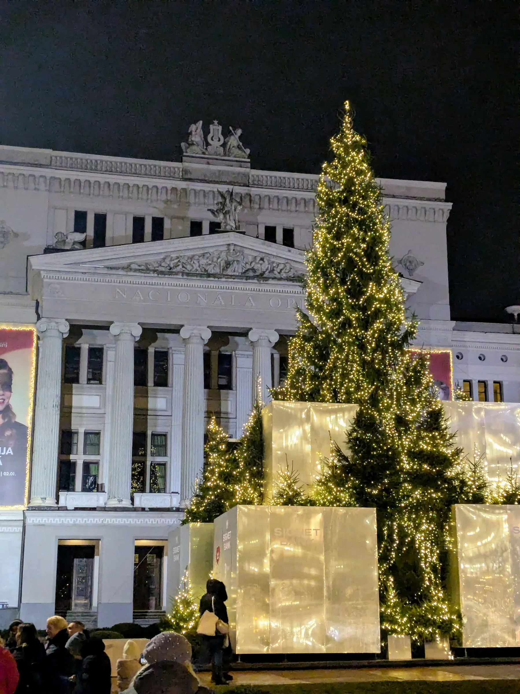 Neoclassical facade of the Latvian National Opera with a monumental illuminated Christmas tree and glowing gift boxes in the evening