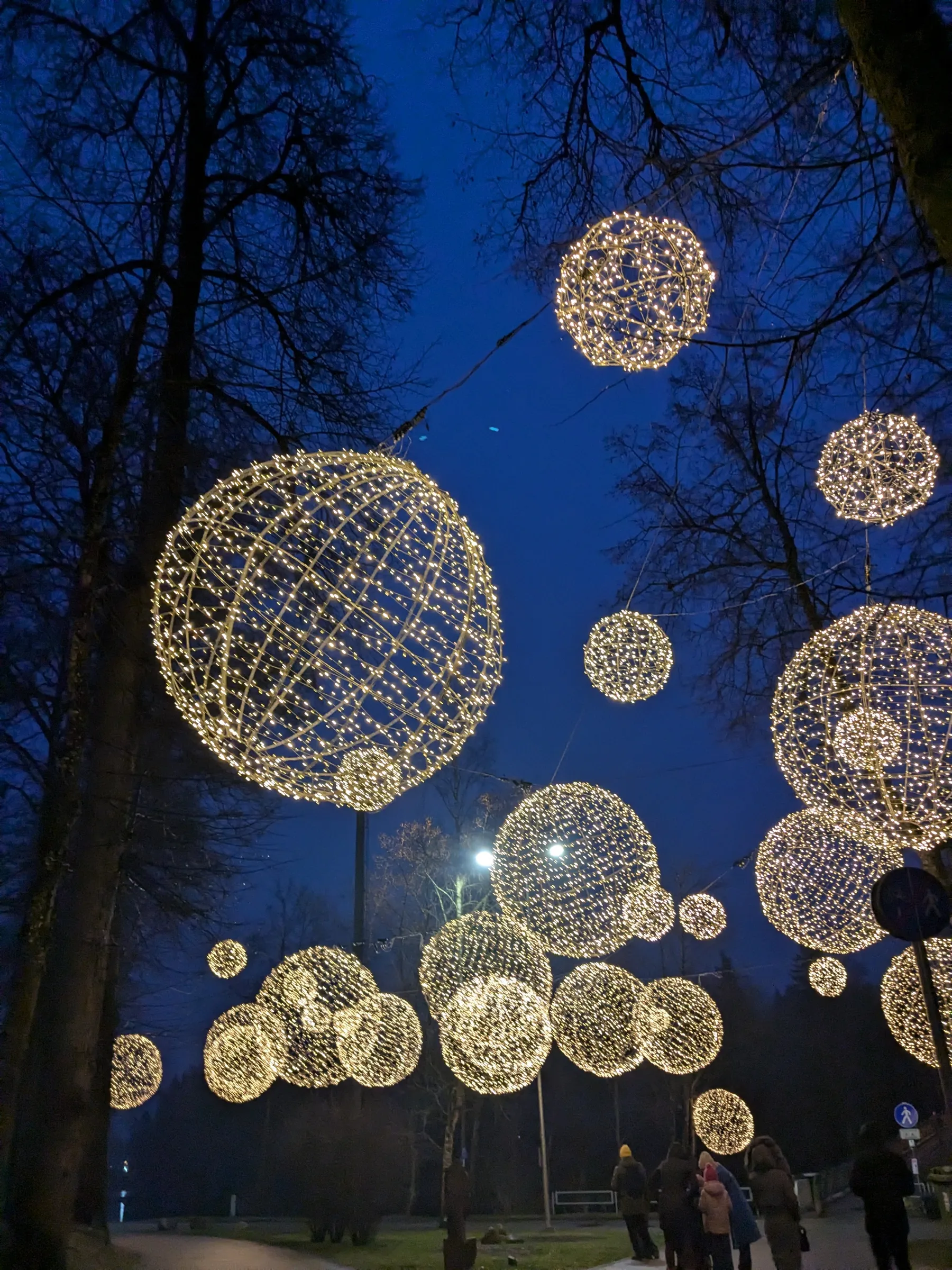 Giant glowing spherical light globes suspended overhead between bare trees above a pedestrian promenade in Ogre at twilight
