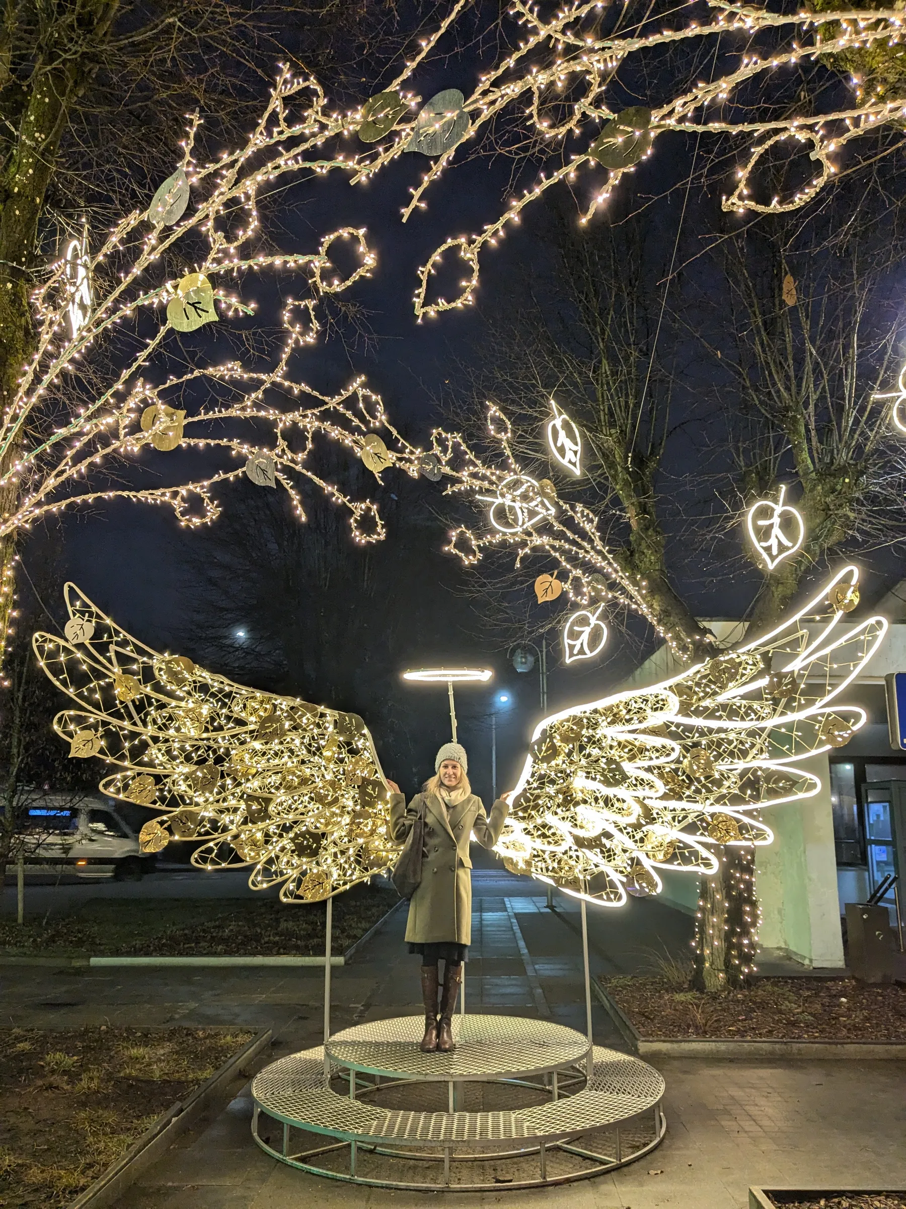 The author standing in front of an illuminated golden angel wings light installation with a glowing halo along Ogre promenade