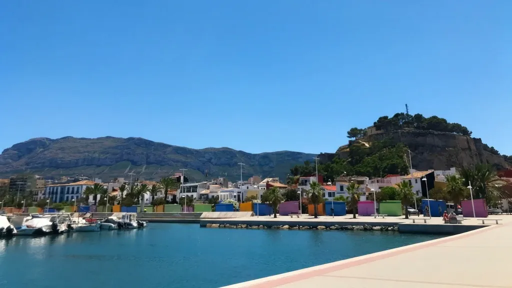 Dénia port with colorful waterfront huts, Castillo de Dénia atop the rocky hill, and the Montgó mountain ridge in the background