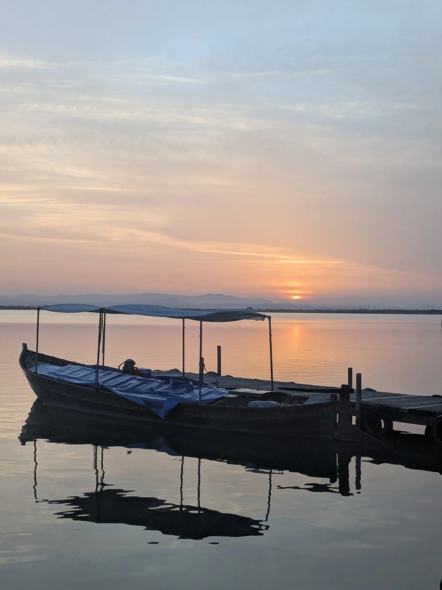 Traditional flat-bottom wooden boat moored at a rustic jetty on the calm waters of Albufera lagoon during sunset