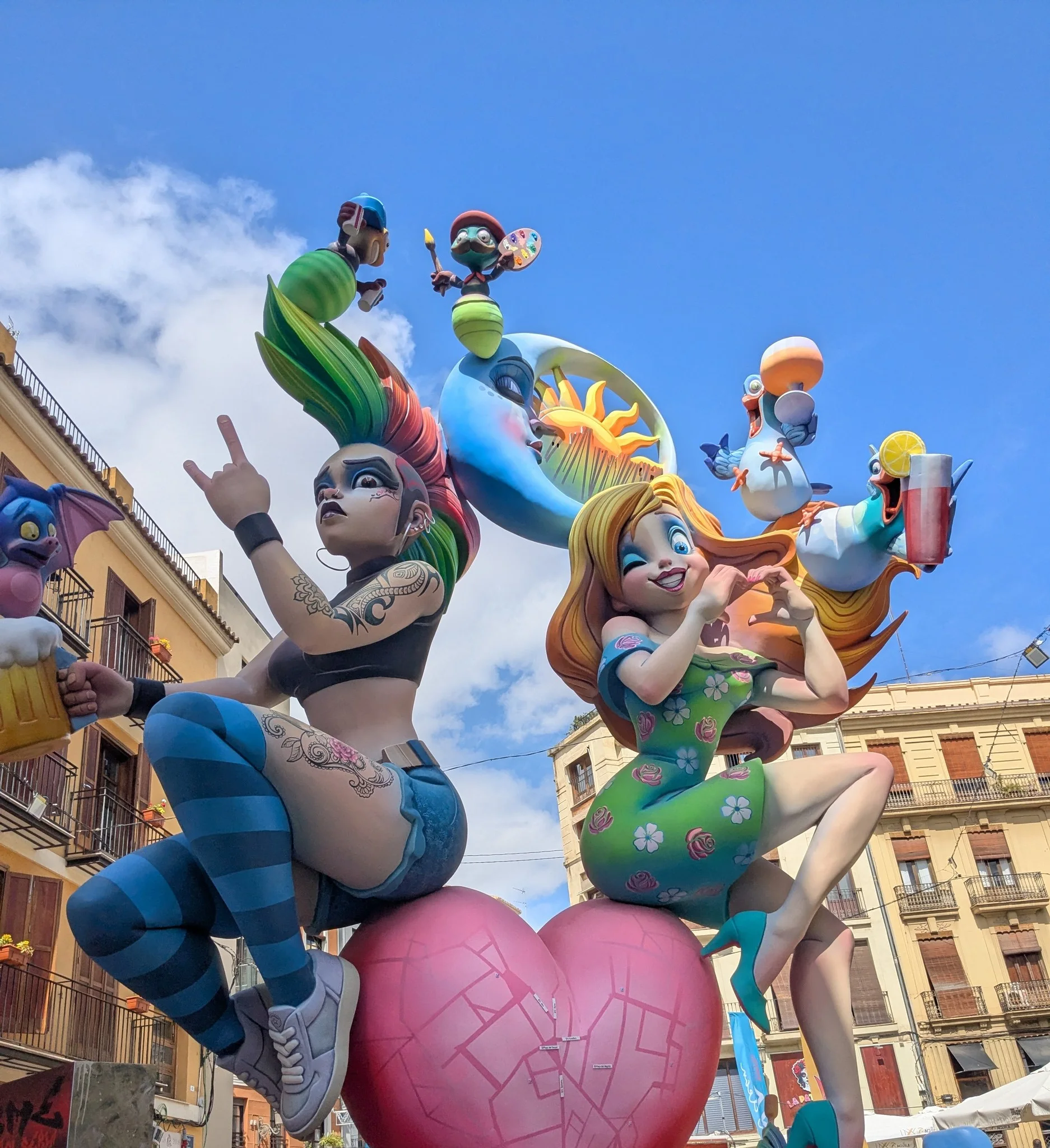 Giant colorful Fallas street sculpture with whimsical punk and floral characters perched on a pink heart under blue sky in Valencia