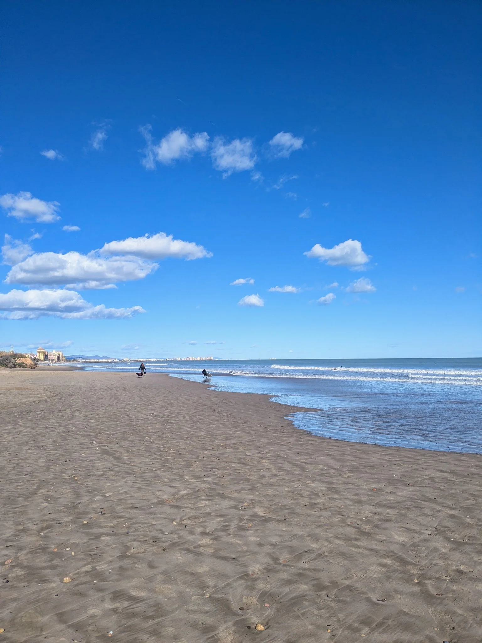 Expansive sandy beach with gentle waves and walking figures along the shoreline at Playa de la Patacona under a bright blue sky